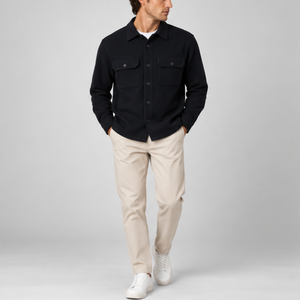 A man wearing a black long-sleeve button-up shirt, beige trousers, and white sneakers, standing against a light gray studio background.